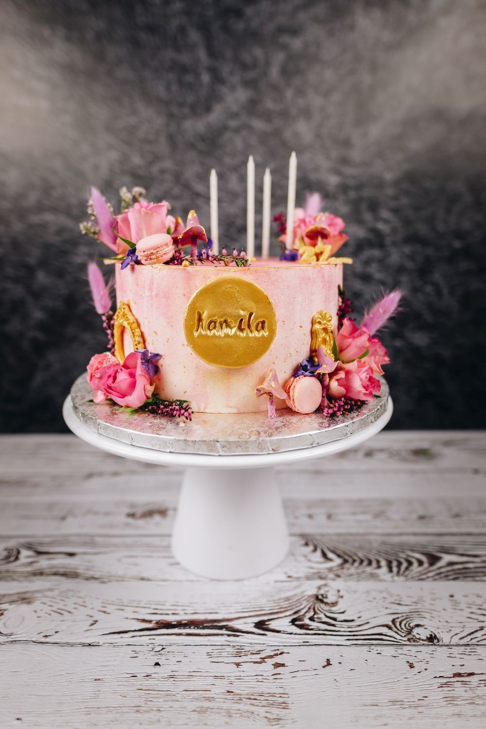 Tort urodzinowy w odcieniach różu, zdobiony świeżymi różami, makaronikami i złotymi akcentami, z napisem 'Kamila' na złotym medalionie, umieszczony na białym stojaku na tle ciemnej ściany z efektem...
