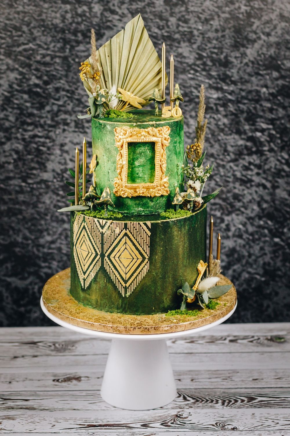 Dwupoziomowy tort w odcieniach zieleni i złota, zdobiony geometrycznymi wzorami, suszonymi roślinami, złotą ramką i świeczkami, umieszczony na białym stojaku.