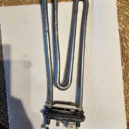 Wyjęta grzałka pralki z widocznymi śladami użytkowania, leżąca na białym ręczniku papierowym na tle plecionej podkładki.