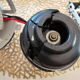 Uszkodzony silnik elektryczny z pękniętą obudową, prawdopodobnie zdemontowany z urządzenia AGD, leży na białym blacie z dekoracyjną, złotą podkładką.