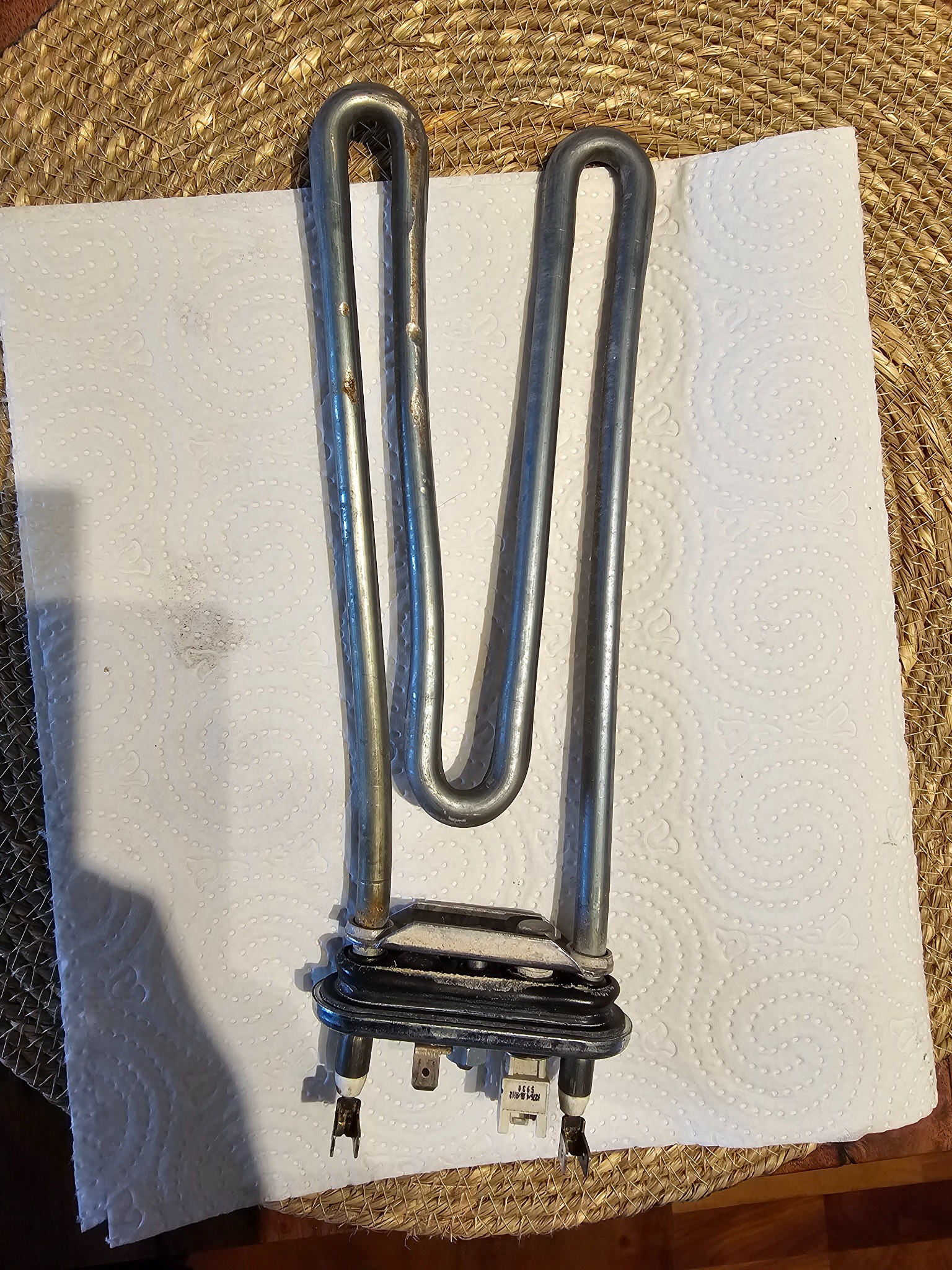 Wyjęta grzałka pralki z widocznymi śladami użytkowania, leżąca na białym ręczniku papierowym na tle plecionej podkładki.