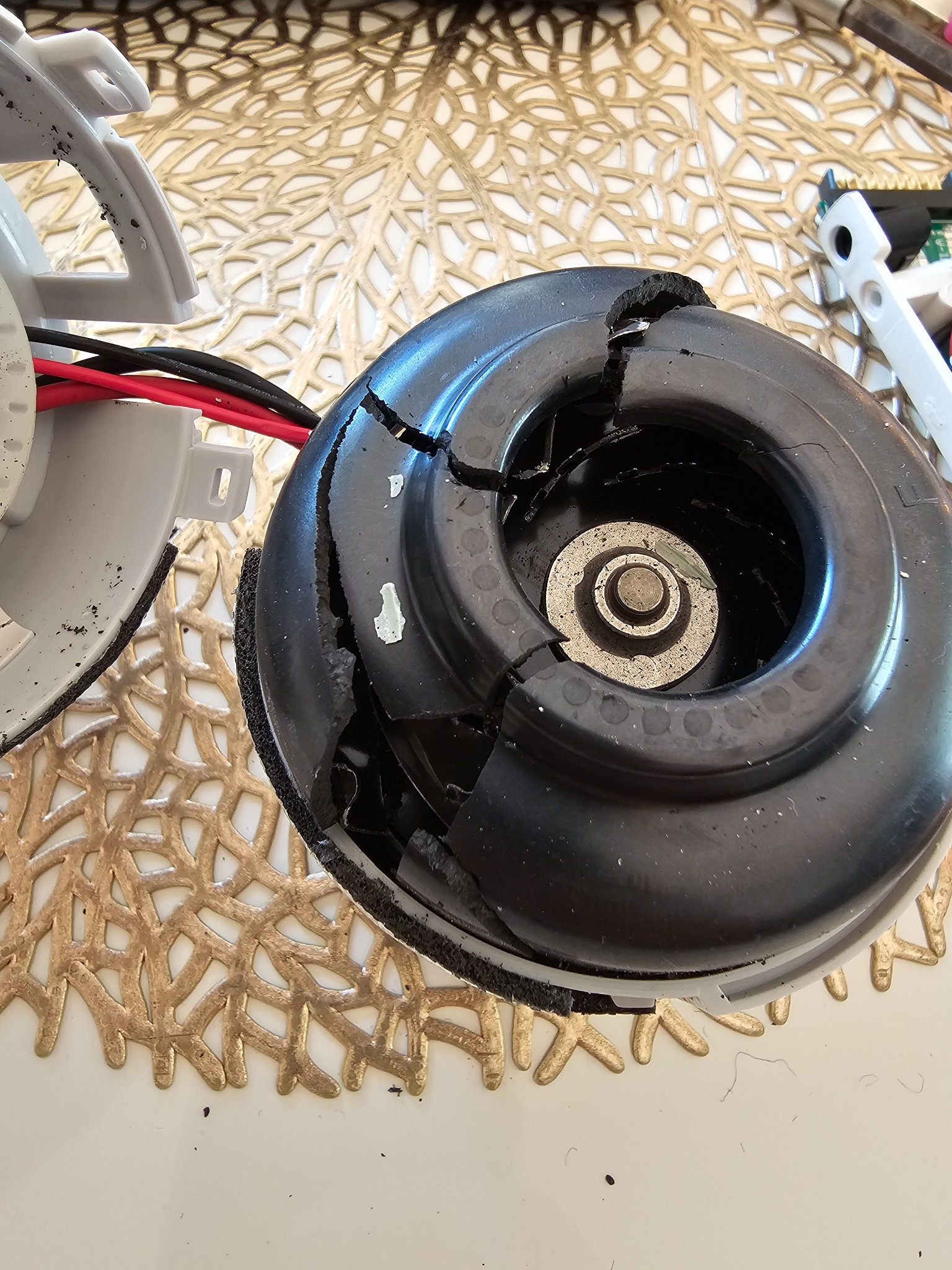 Uszkodzony silnik elektryczny z pękniętą obudową, prawdopodobnie zdemontowany z urządzenia AGD, leży na białym blacie z dekoracyjną, złotą podkładką.