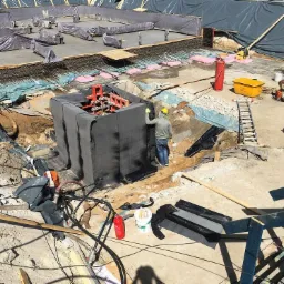 Budowa fundament&oacute;w: pracownik w kasku przy wykopie z zabezpieczeniem, widoczne zbrojenia, betonowe elementy, folie ochronne i sprzęt budowlany.