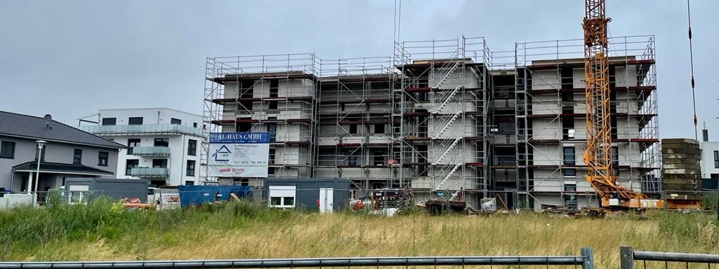 Wielopiętrowy budynek w stanie surowym otwartym, z rusztowaniami i dźwigiem w tle, na pierwszym planie trawa i ogrodzenie.