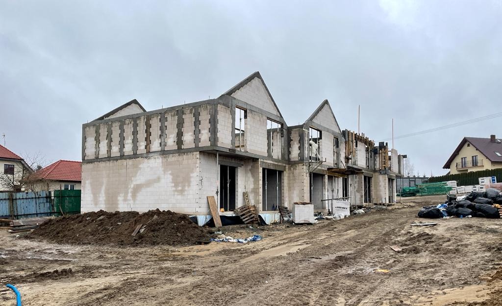 Stan surowy zamknięty szeregu domów jednorodzinnych z betonu komórkowego, widoczne ślady po szalunkach na wieńcu, teren budowy w trakcie prac ziemnych, pochmurne niebo.