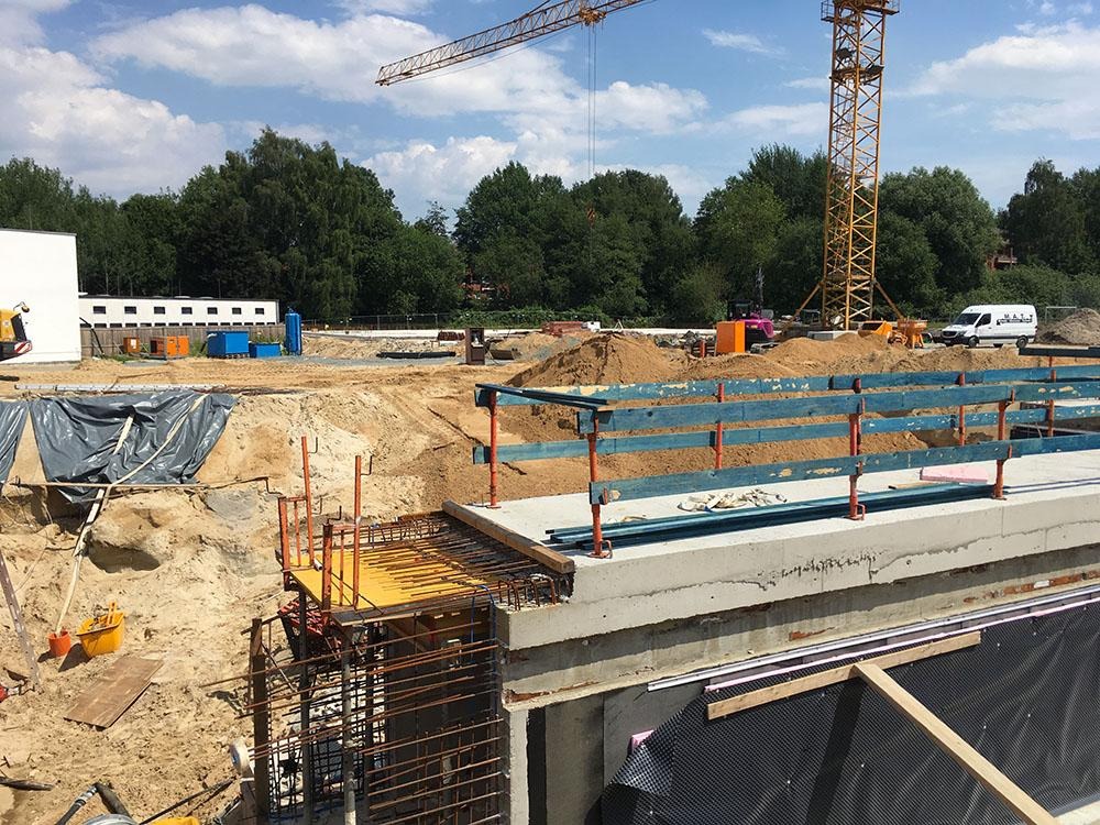 Budowa fundamentów z widocznym zbrojeniem, deskowaniem, oraz zabezpieczeniem przeciwwilgociowym, na tle placu budowy z dźwigami i piaskiem.