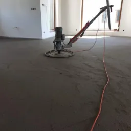 Zacieraczka mechaniczna do jastrychu w trakcie pracy na świeżo wylanym, szarym jastrychu w jasnym pomieszczeniu z dwoma oknami.