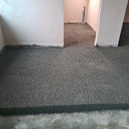 P&G POSADZKI - Szara, świeżo wylana posadzka jastrychowa w pomieszczeniu z widocznymi progami drzwiowymi. W tle fragment instalacji grzewczej.