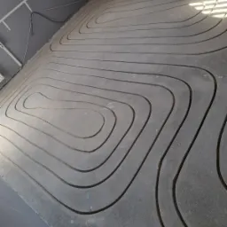 Szara, betonowa wylewka z wyfrezowanymi rowkami pod instalację grzewczą. Widok z góry na podłogę w trakcie prac instalacyjnych.