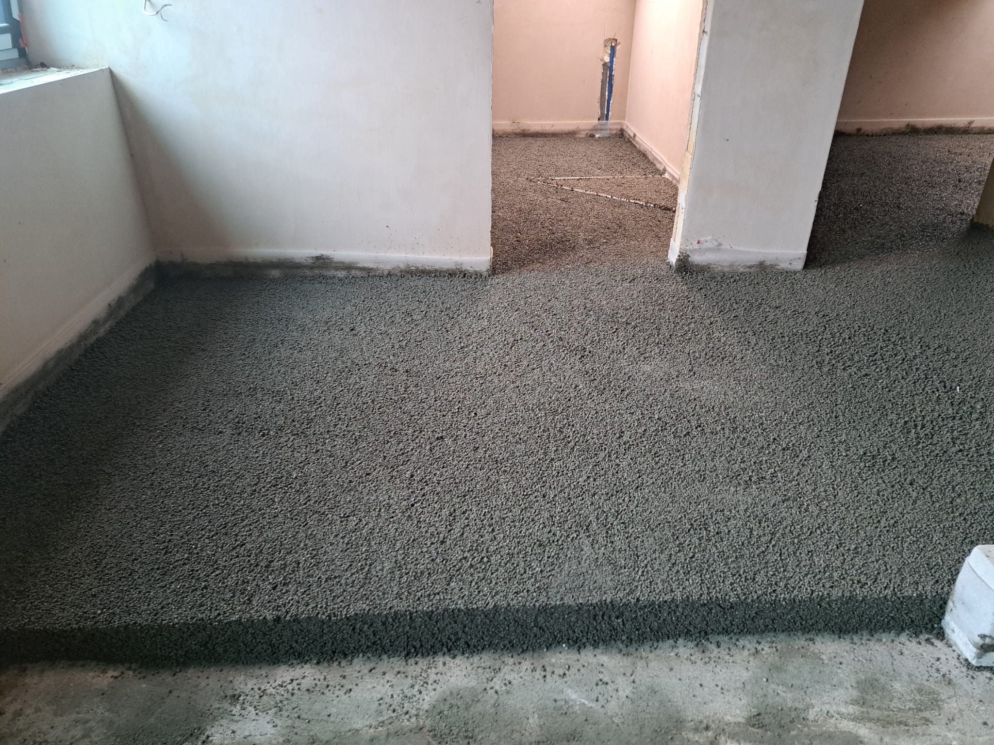 Szara, świeżo wylana posadzka jastrychowa w pomieszczeniu z widocznymi progami drzwiowymi. W tle fragment instalacji grzewczej.