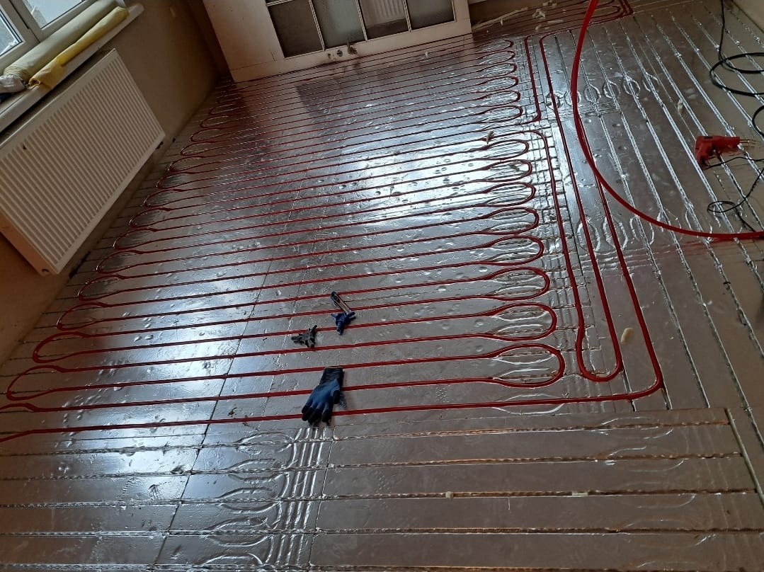 Czerwone rury ogrzewania podłogowego ułożone na srebrnej izolacji, z narzędziami i rękawicą roboczą. Widok z góry na instalację w trakcie montażu.