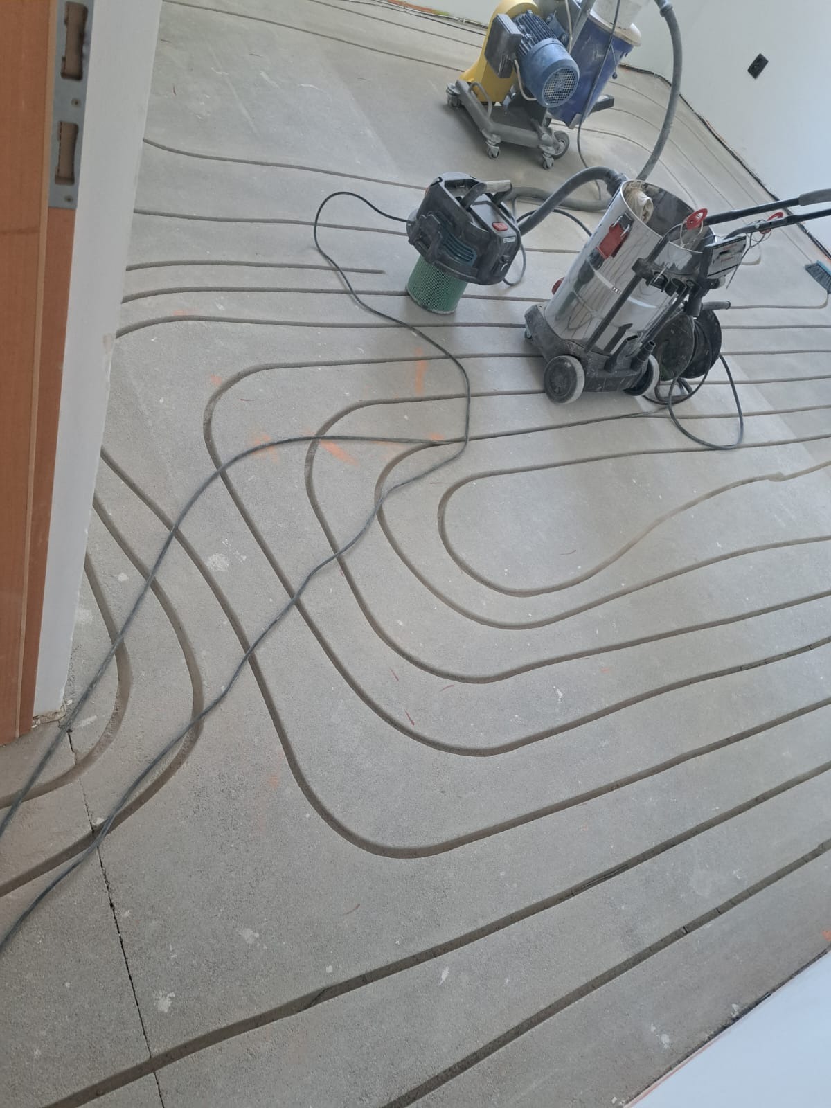 Szlifowana posadzka jastrychowa z frezowanymi kanałami pod rury ogrzewania podłogowego, widoczne narzędzia: szlifierka i odkurzacz przemysłowy.