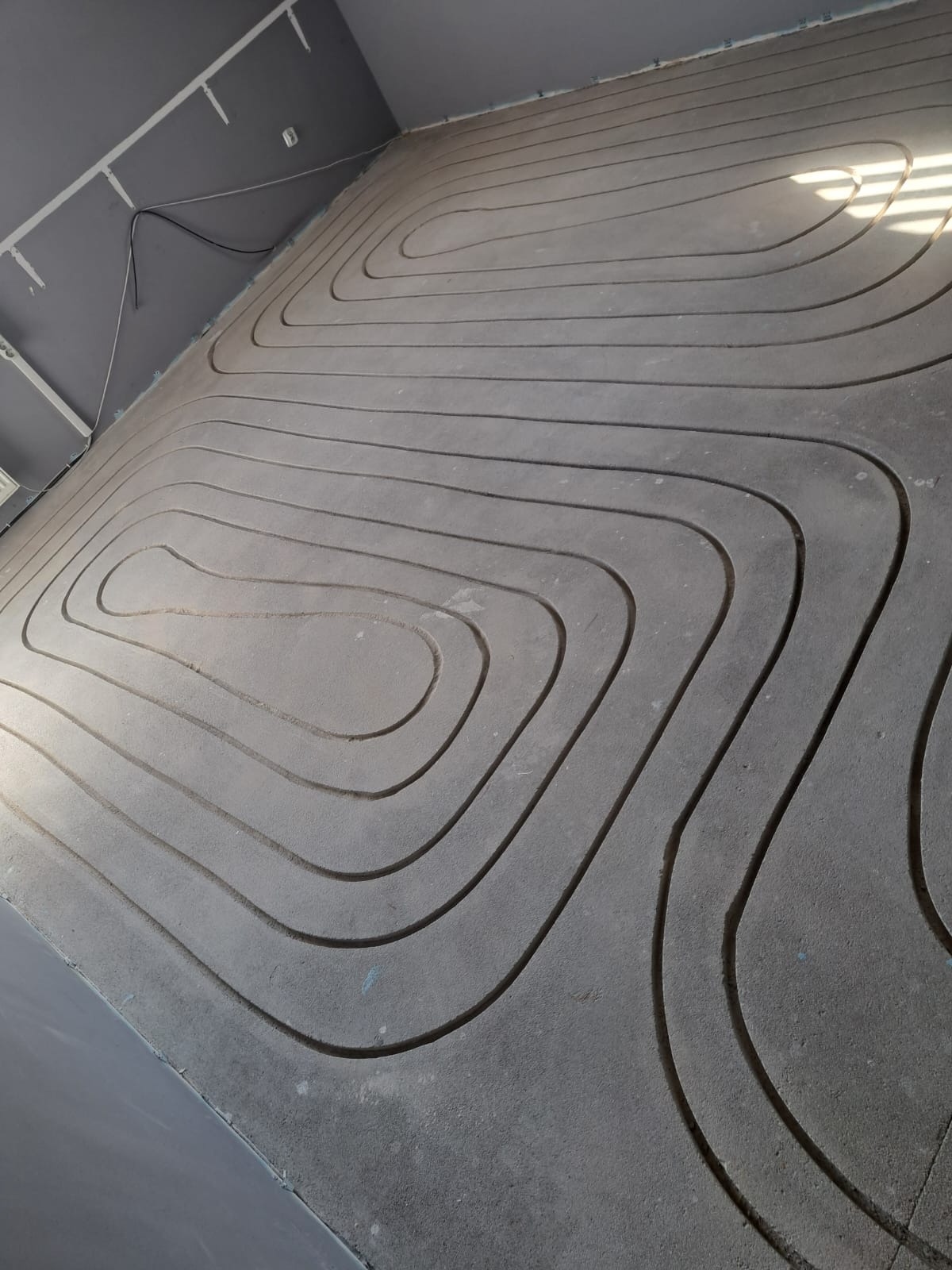 Szara, betonowa wylewka z wyfrezowanymi rowkami pod instalację grzewczą. Widok z góry na podłogę w trakcie prac instalacyjnych.