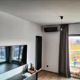 Czarny klimatyzator ścienny zamontowany w nowoczesnym salonie z widokiem na plac zabaw przez okno, obok czarny grzejnik panelowy.