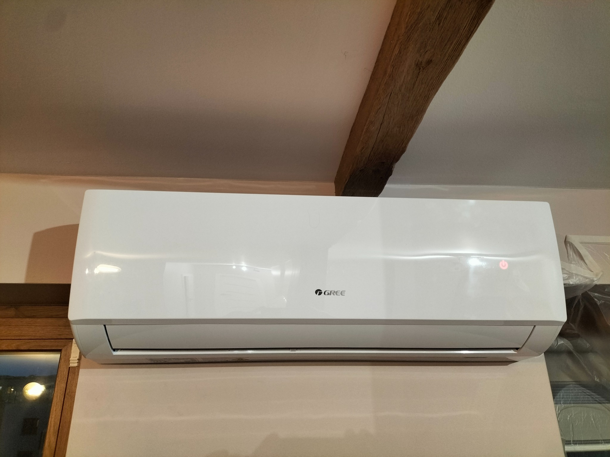 Biała jednostka klimatyzacyjna marki Gree zamontowana pod sufitem z widoczną drewnianą belką stropową, w tle okno.