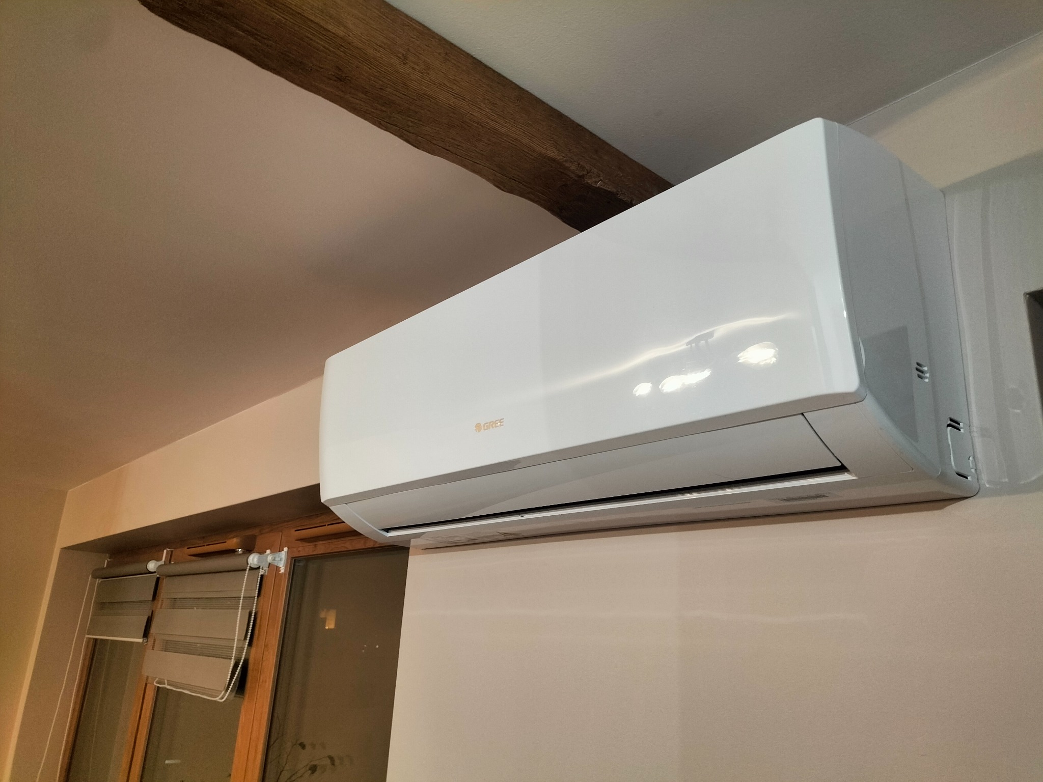 Biały klimatyzator marki Gree zamontowany na ścianie w pomieszczeniu z drewnianą belką stropową i oknem z roletami.