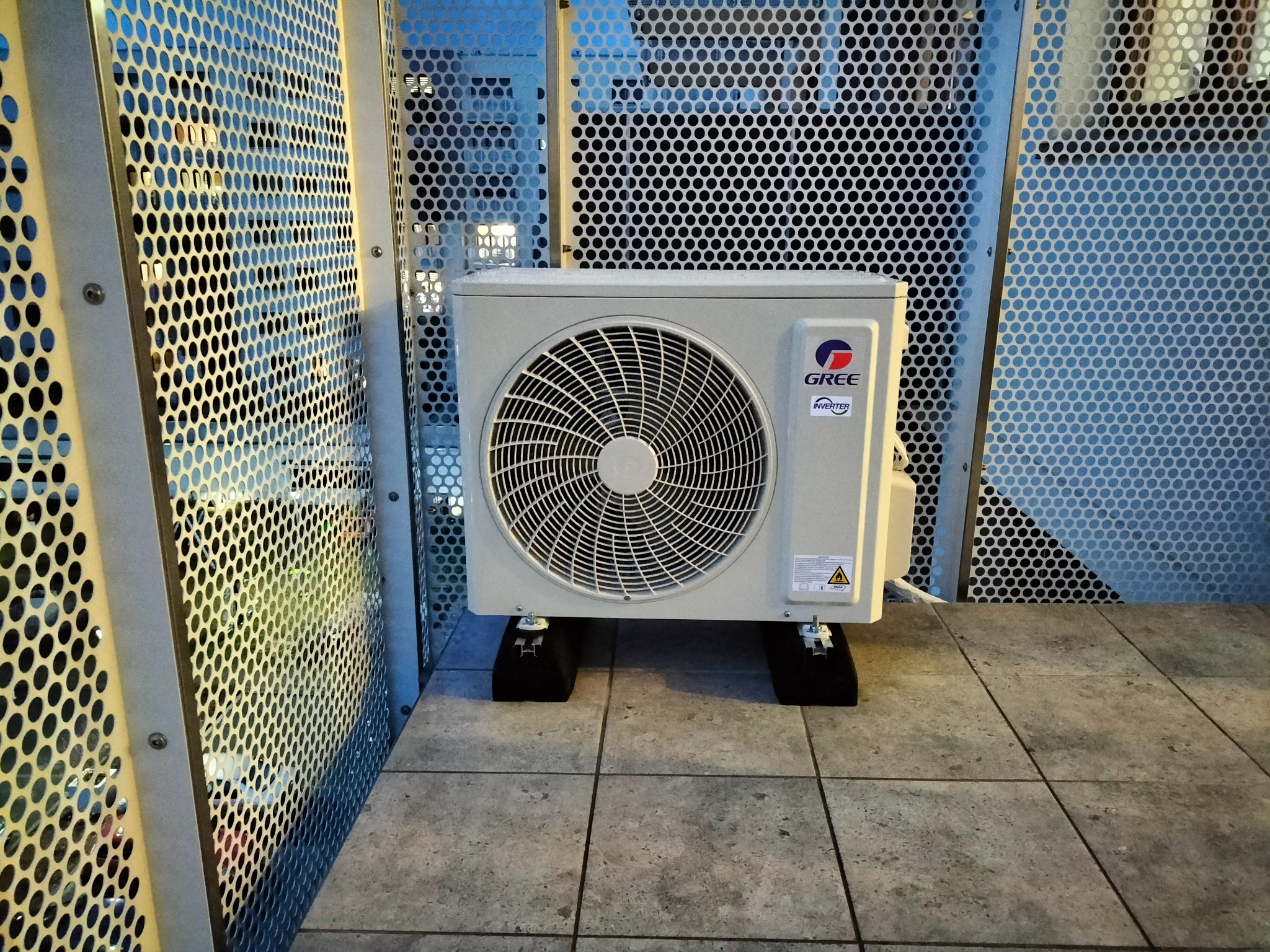 Zewnętrzna jednostka klimatyzacji Gree Inverter umieszczona na czarnych podkładkach antywibracyjnych, otoczona metalową perforowaną klatką ochronną na szarej, kwadratowej posadzce.
