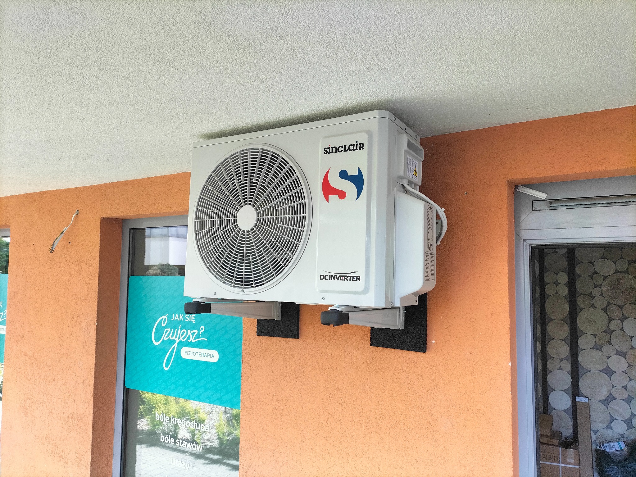 Zewnętrzna jednostka klimatyzacji Sinclair DC Inverter zamontowana na pomarańczowej fasadzie budynku z widocznym logo i naklejką ostrzegawczą, zainstalowana na metalowych wspornikach z czarnymi...