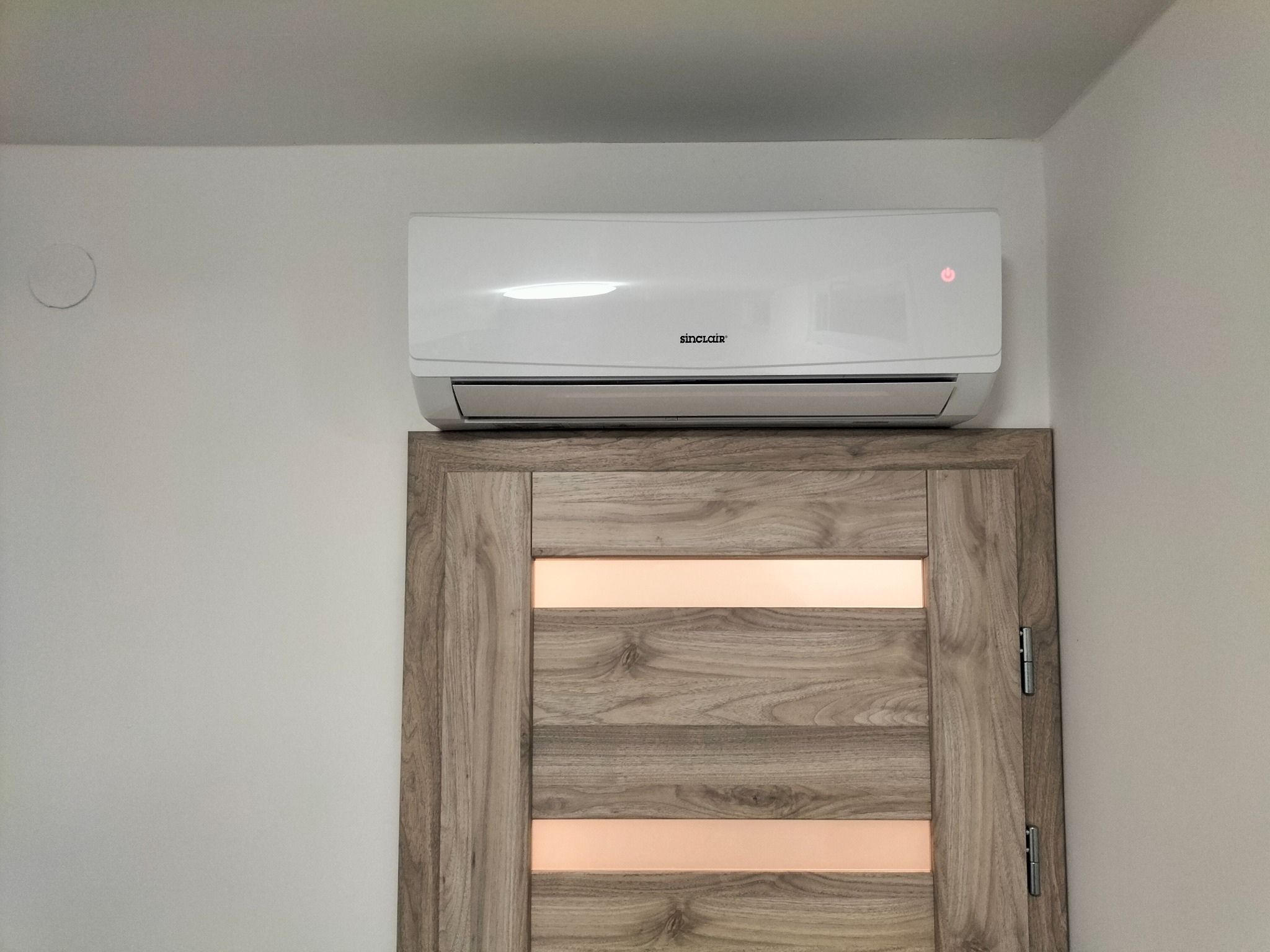 Biały klimatyzator Sinclair zamontowany nad drewnianymi drzwiami z poziomymi przeszkleniami, widoczna dioda sygnalizacyjna pracy urządzenia.