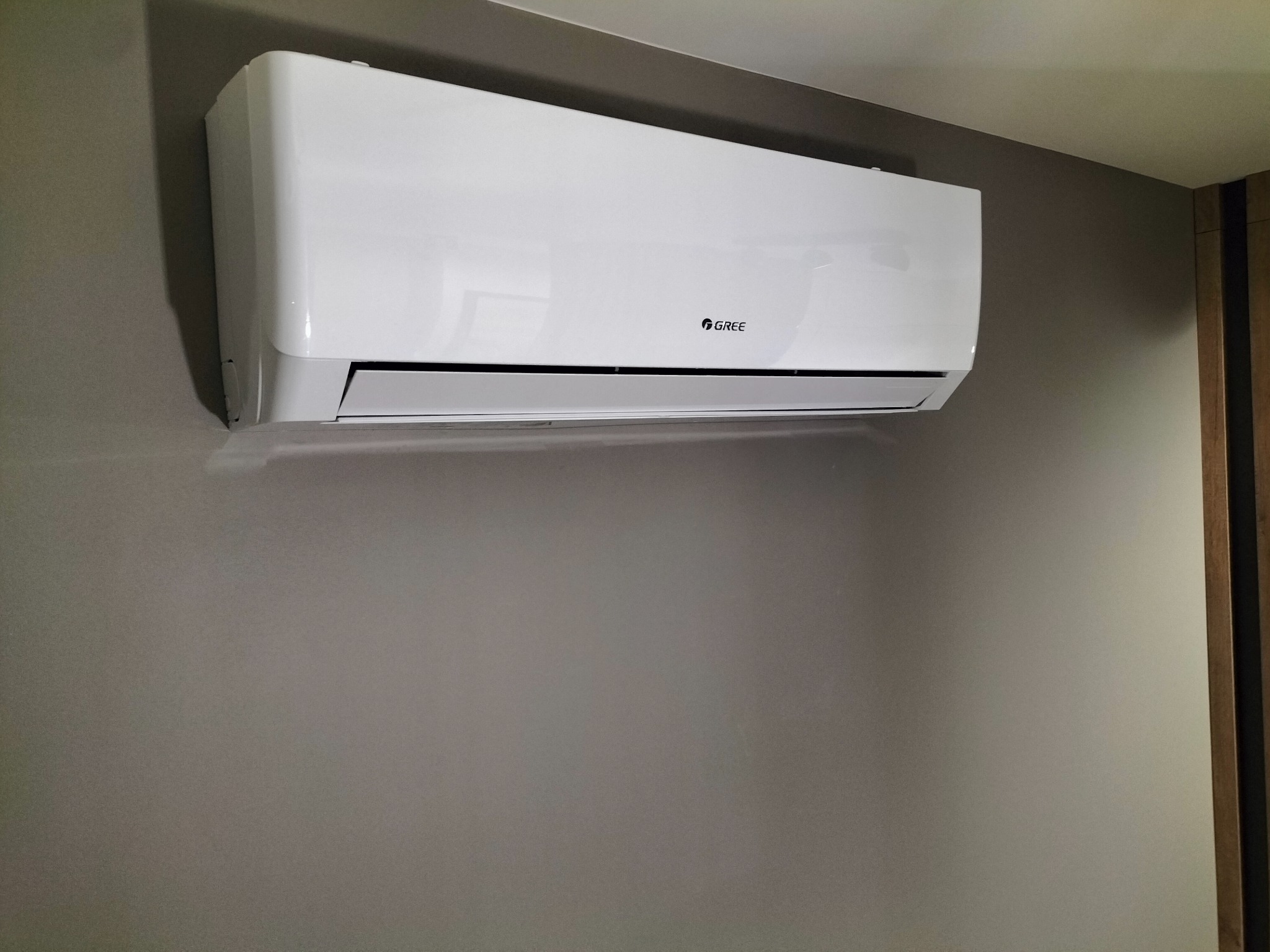 Biała klimatyzacja marki Gree zamontowana na szarej ścianie w pomieszczeniu, widoczny fragment drewnianej ramy drzwi.