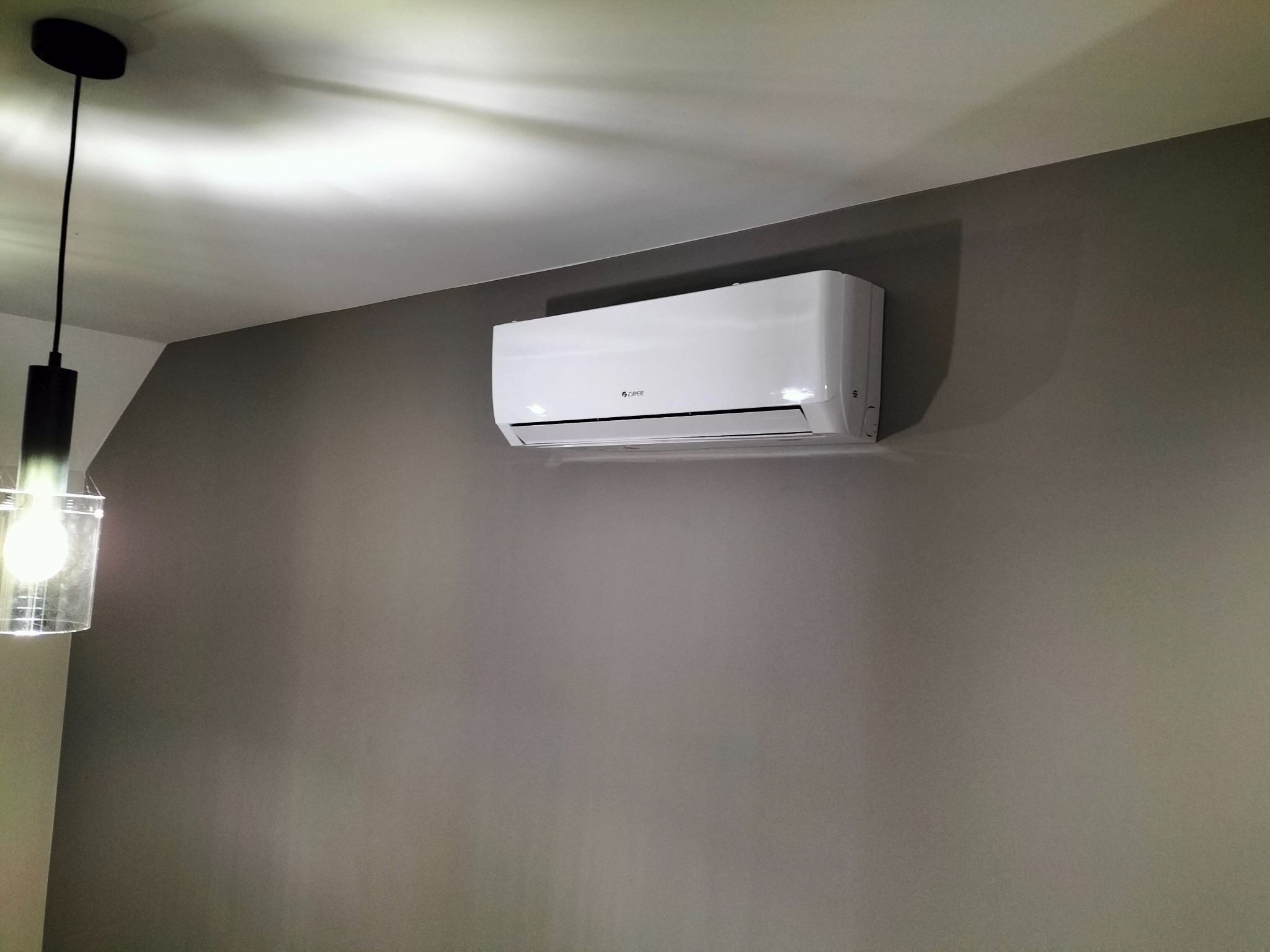 Biały klimatyzator marki Gree zamontowany na szarej ścianie w pokoju, widoczna nowoczesna lampa z żarówką w lewym górnym rogu.
