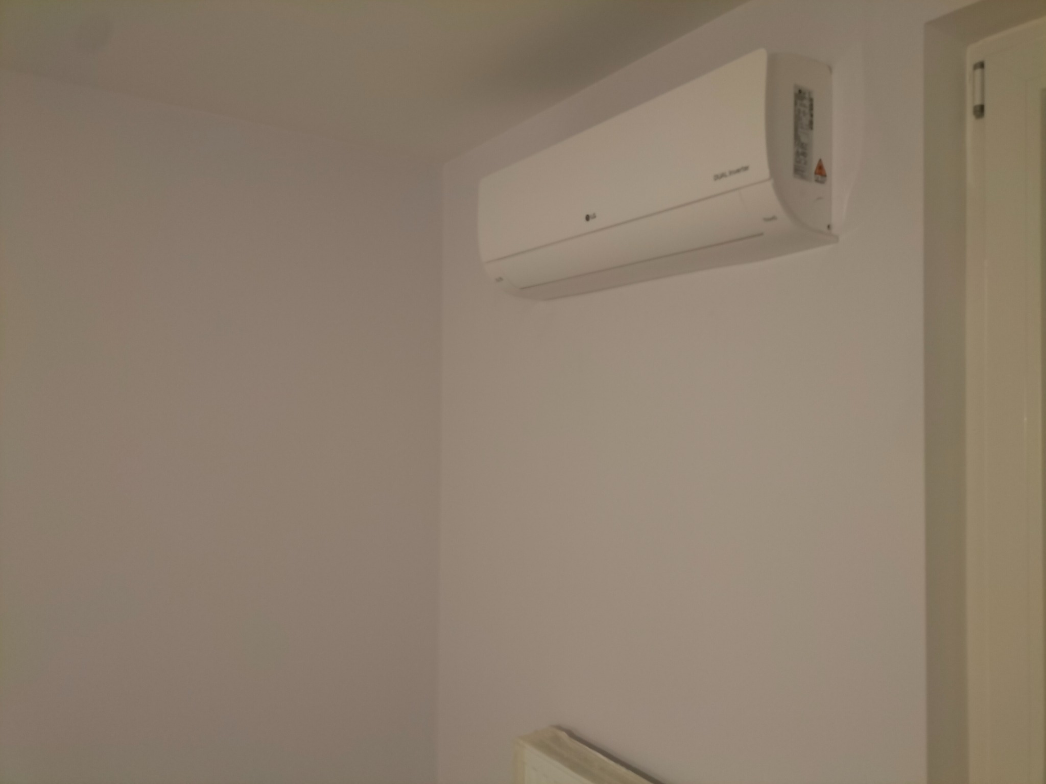 Biała jednostka klimatyzacji LG Dual Inverter zamontowana na ścianie w pomieszczeniu, widoczna obok fragmentu białych drzwi.