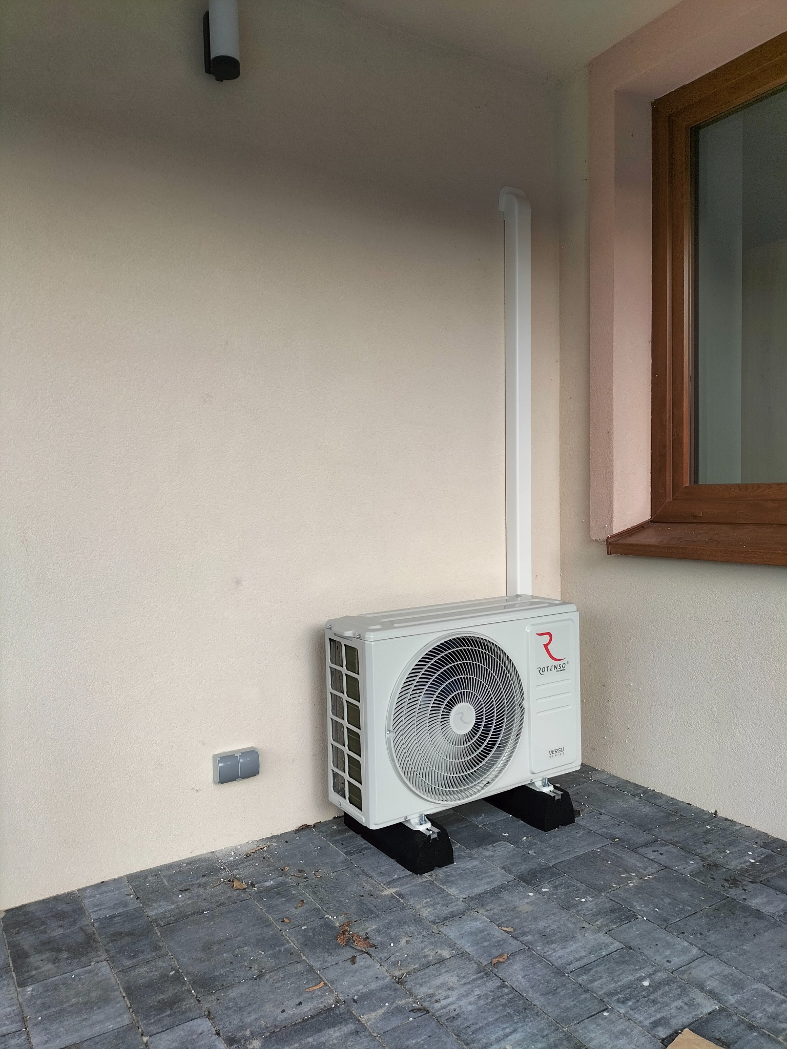 Zewnętrzna jednostka klimatyzacji ROTENSO zamontowana na czarnych podkładkach antywibracyjnych przy jasnej ścianie budynku z widocznym oknem w drewnianej ramie i rurą odprowadzającą skropliny.