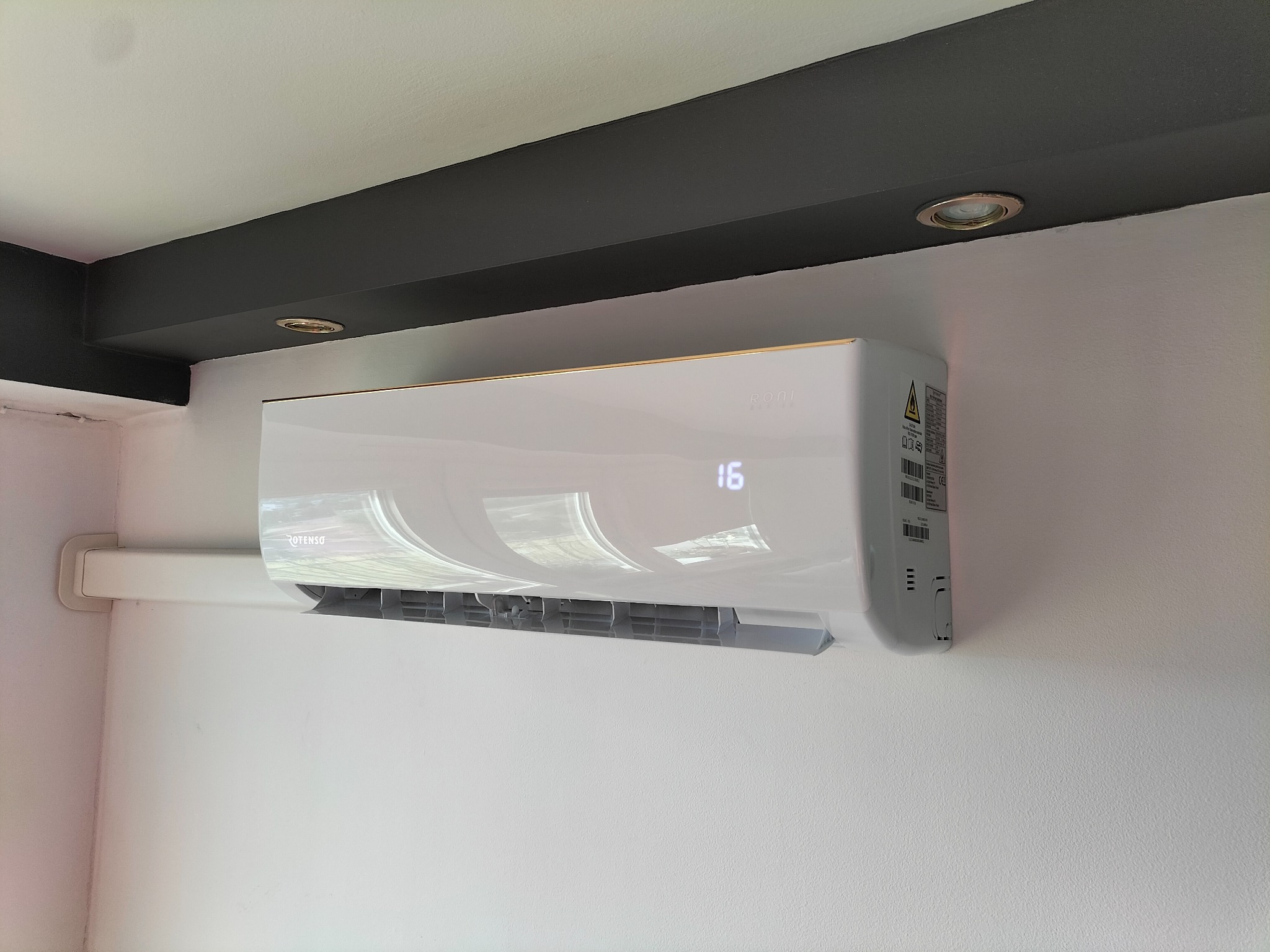 Biała klimatyzacja ścienna z wyświetlaczem temperatury (16 stopni) zamontowana w pomieszczeniu z ciemną belką sufitową i oświetleniem punktowym.