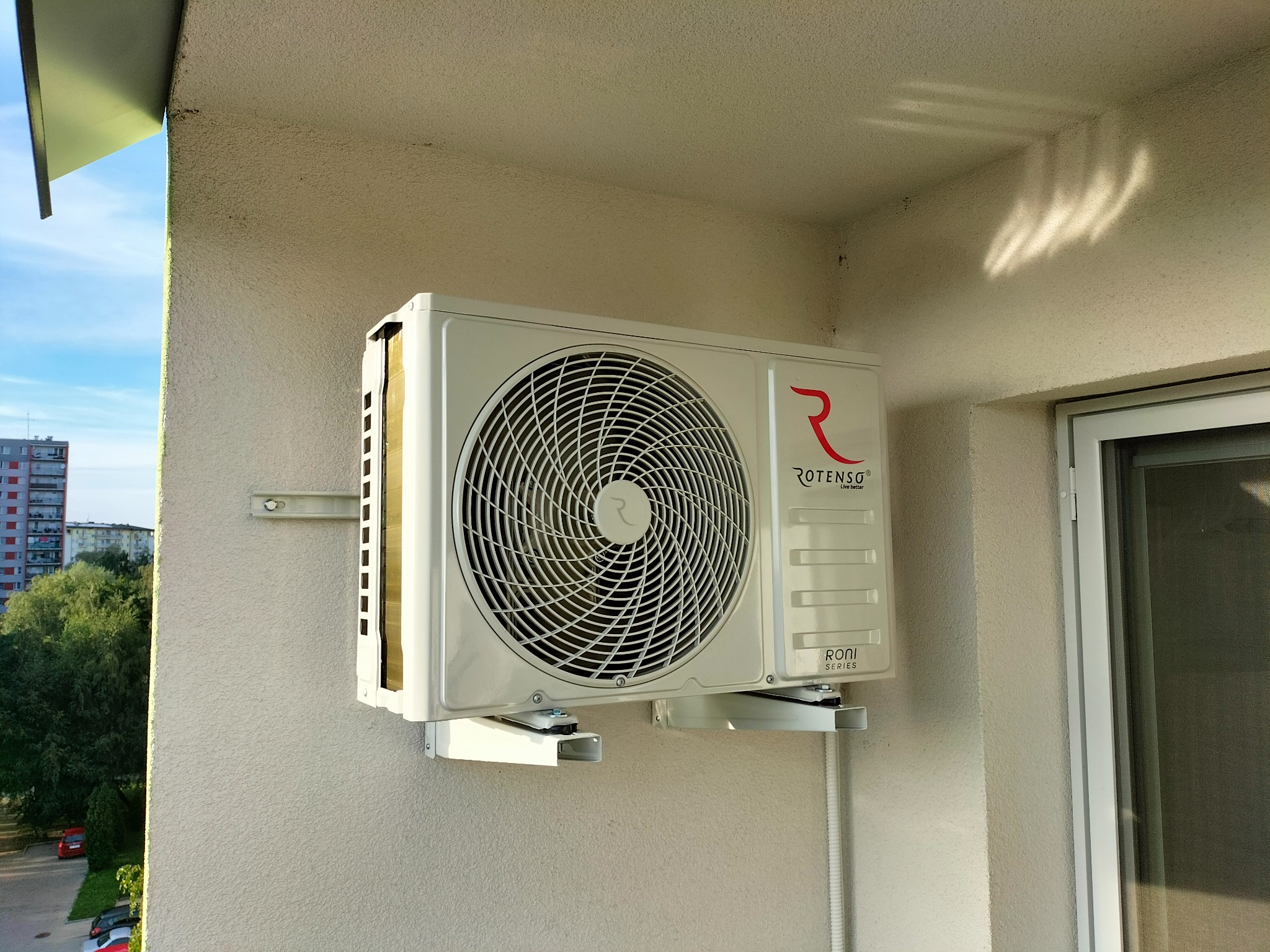 Biała jednostka zewnętrzna klimatyzacji Rotenso Roni Series zamontowana na balkonie mieszkania w bloku; widok z boku na elewację budynku i zieleń.