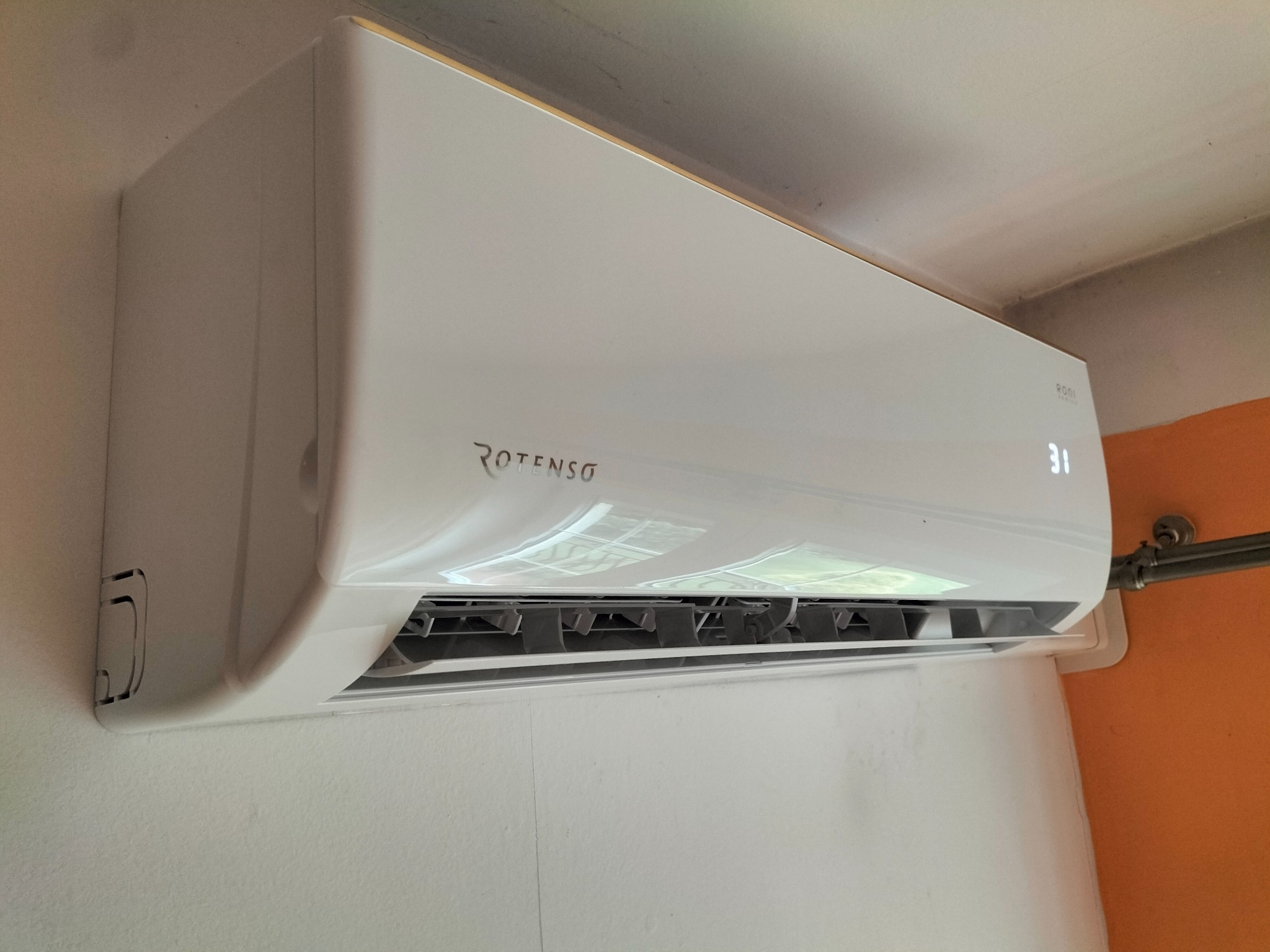 Biały klimatyzator Rotenso zamontowany na ścianie, wyświetlający temperaturę 31 stopni Celsjusza, widoczny fragment instalacji rurowej.