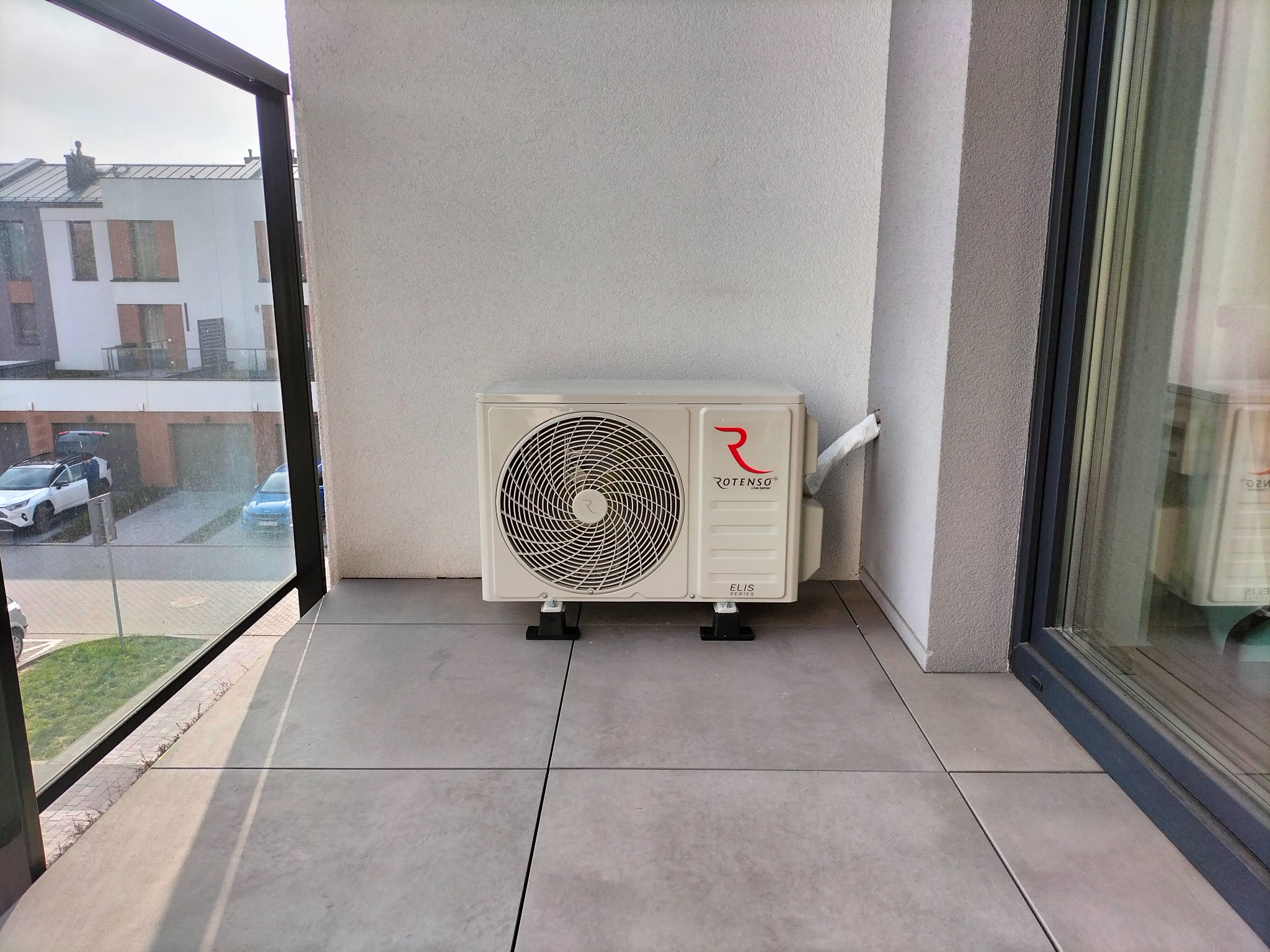 Kompaktowa jednostka zewnętrzna klimatyzacji Rotenso Elis Series zainstalowana na balkonie w Michałowicach, widok z bliska na szarych płytkach podłogowych, z widokiem na budynki mieszkalne za oknem.