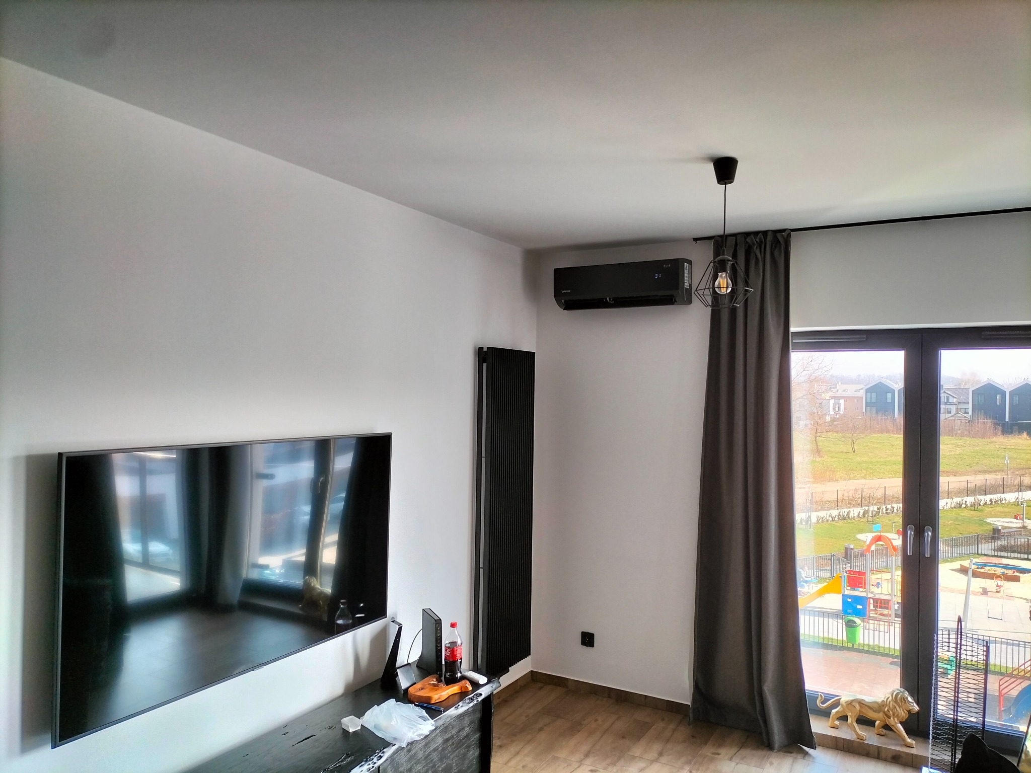 Czarny klimatyzator ścienny zamontowany w nowoczesnym salonie z widokiem na plac zabaw przez okno, obok czarny grzejnik panelowy.