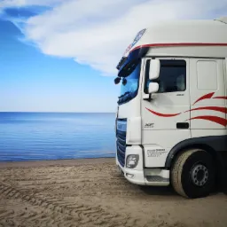 Biała ciężarówka DAF z czerwonymi akcentami, zaparkowana na piaszczystej plaży z widokiem na morze i błękitne niebo z chmurami. Widoczne ślady opon na piasku.