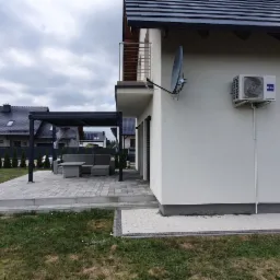 Zewnętrzna jednostka klimatyzacji Haier zamontowana na ścianie domu jednorodzinnego z widoczną instalacją, obok okno z drewnianą ramą i częściowo rozpakowany karton z nowym urządzeniem.