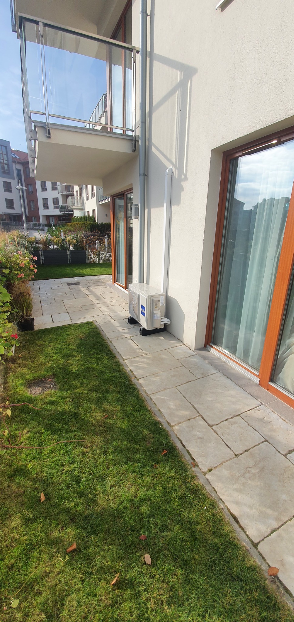 Biała jednostka zewnętrzna klimatyzacji Haier zamontowana na czarnych podstawkach przy ścianie budynku z oknem i balkonem, widoczna trawa i kostka brukowa.