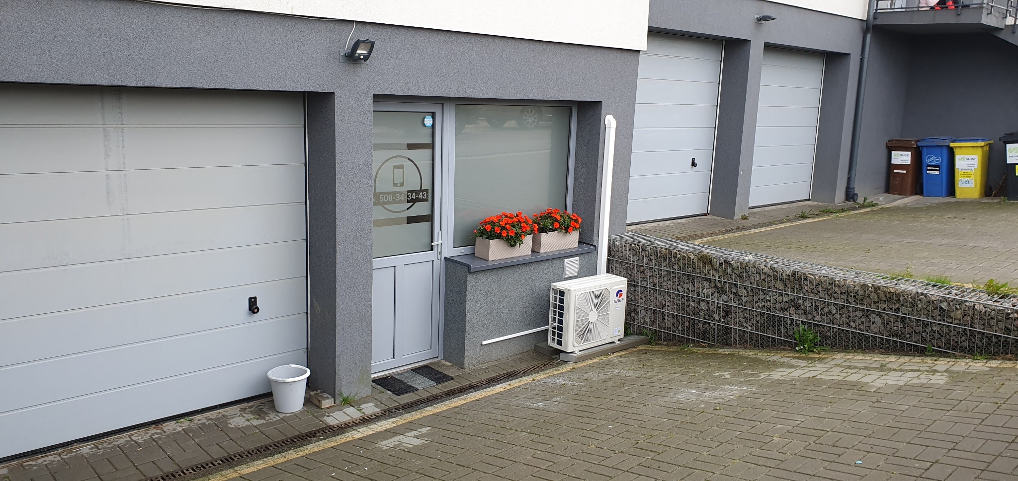 Biała jednostka zewnętrzna klimatyzacji GREE zamontowana na betonowej podstawie przy szarej ścianie budynku z dwoma bramami garażowymi i drzwiami wejściowymi z numerem telefonu na szybie, ozdobiona...