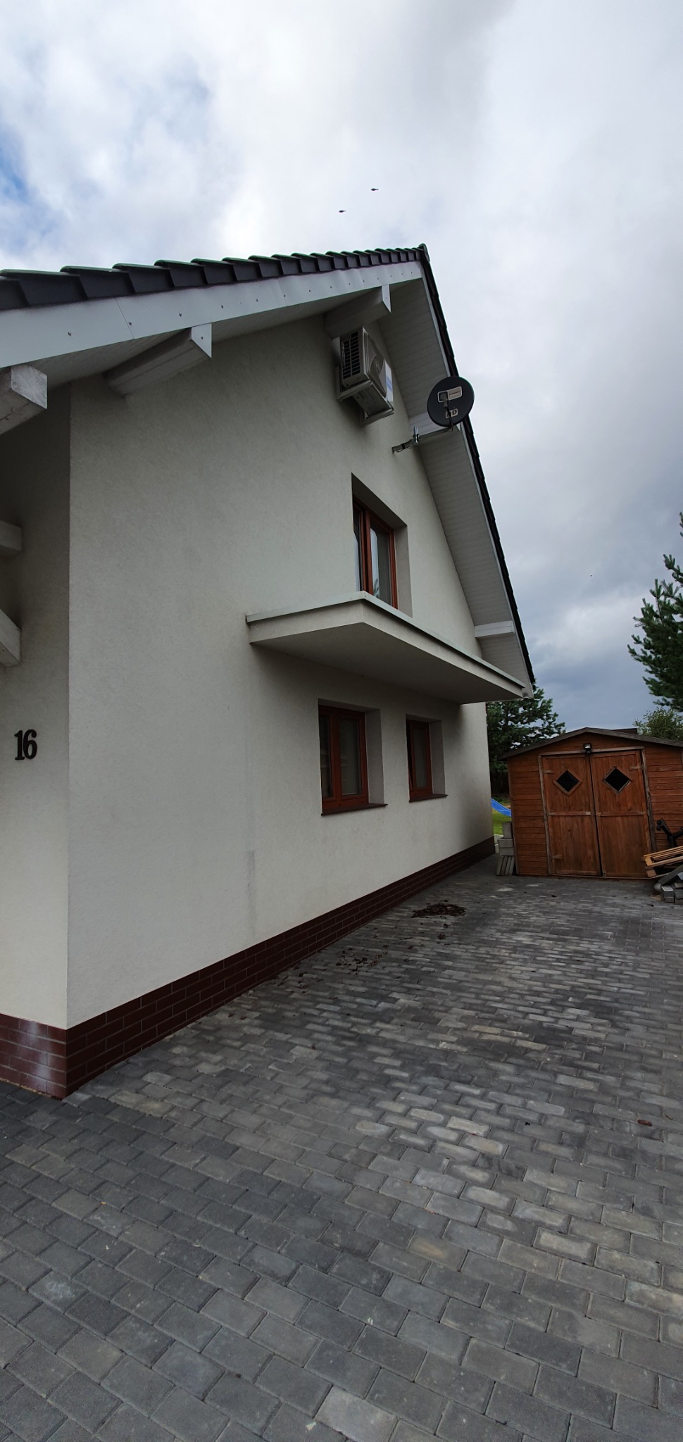 Biały dom jednorodzinny z zamontowaną klimatyzacją na elewacji, widoczne brązowe okna i numer domu 16.
