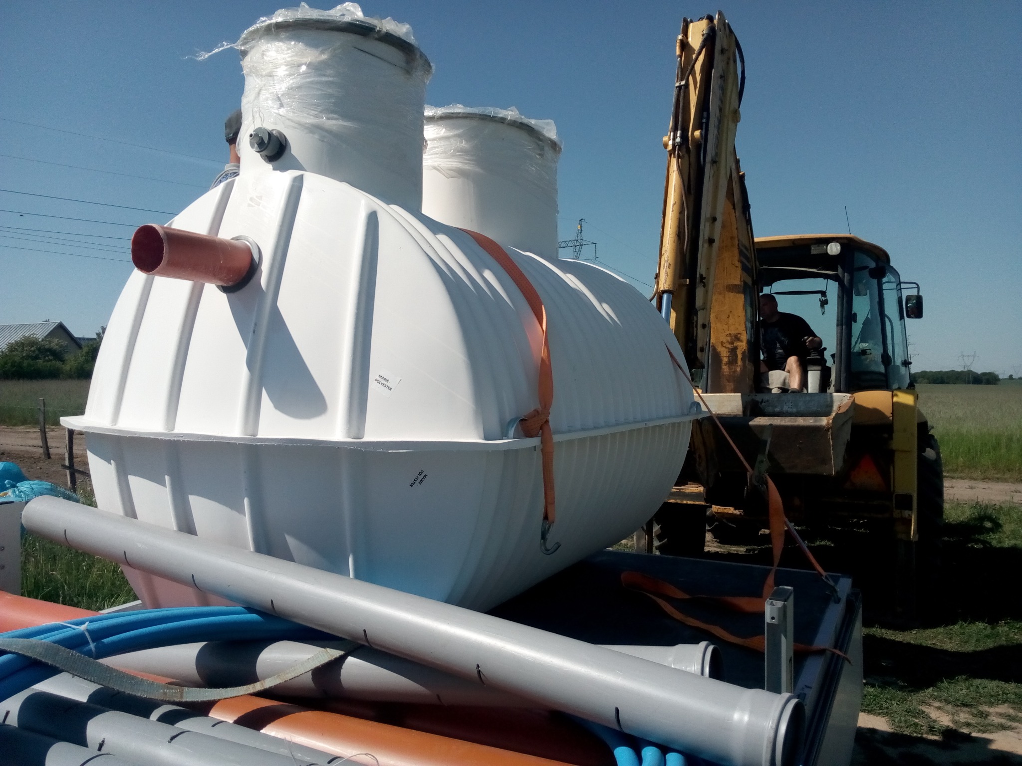 Biały, żebrowany zbiornik z pomarańczowym króćcem, zabezpieczony pasami transportowymi, umieszczony na przyczepie, obok żółtej koparki z operatorem w kabinie, na tle pola i błękitnego nieba.