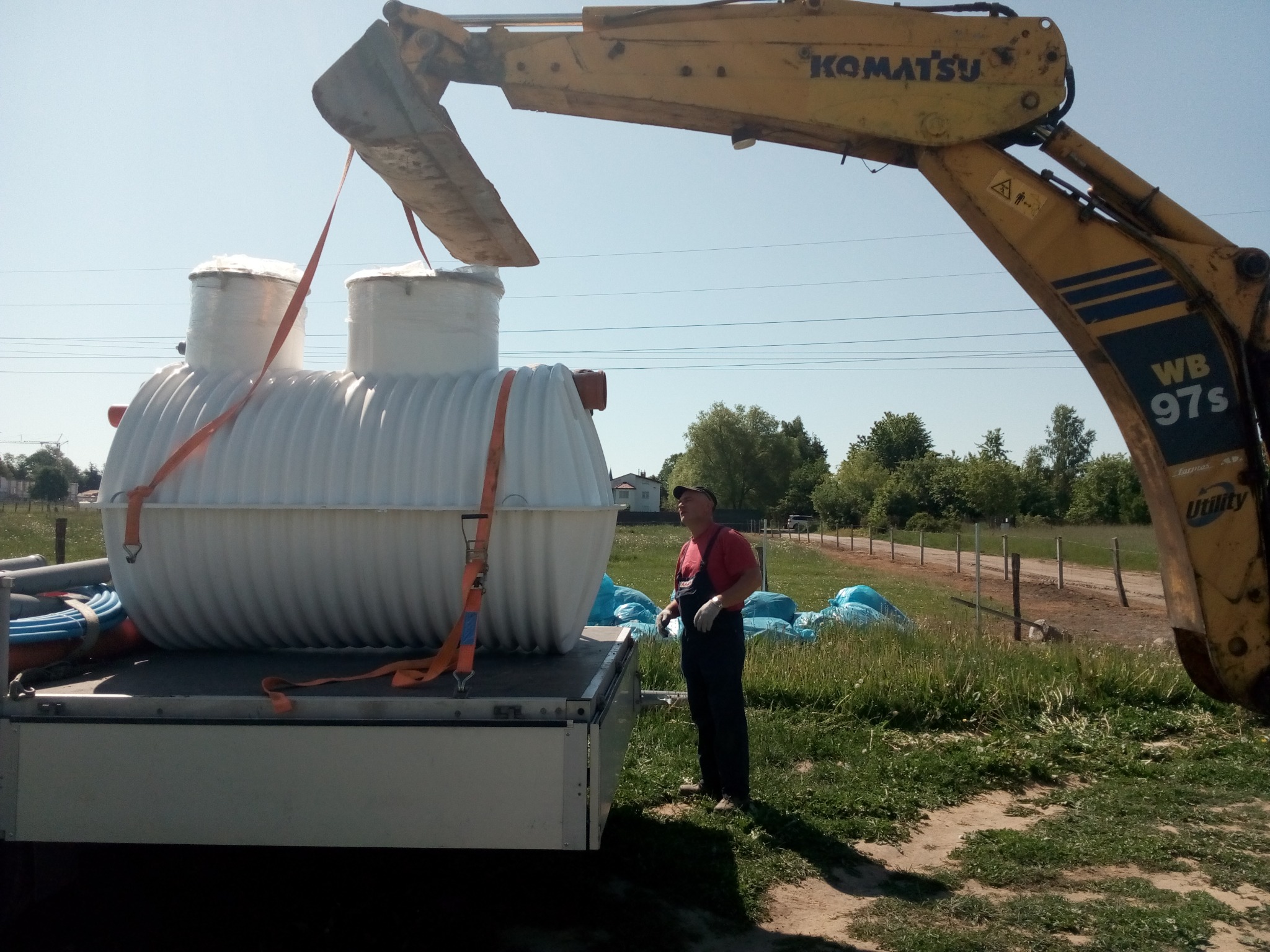 Żółta koparka Komatsu WB 97S podnosi biały, żebrowany zbiornik na ścieki za pomocą pomarańczowych pasów, umieszczając go na platformie ciężarówki; pracownik w czerwonej koszulce i czarnych...