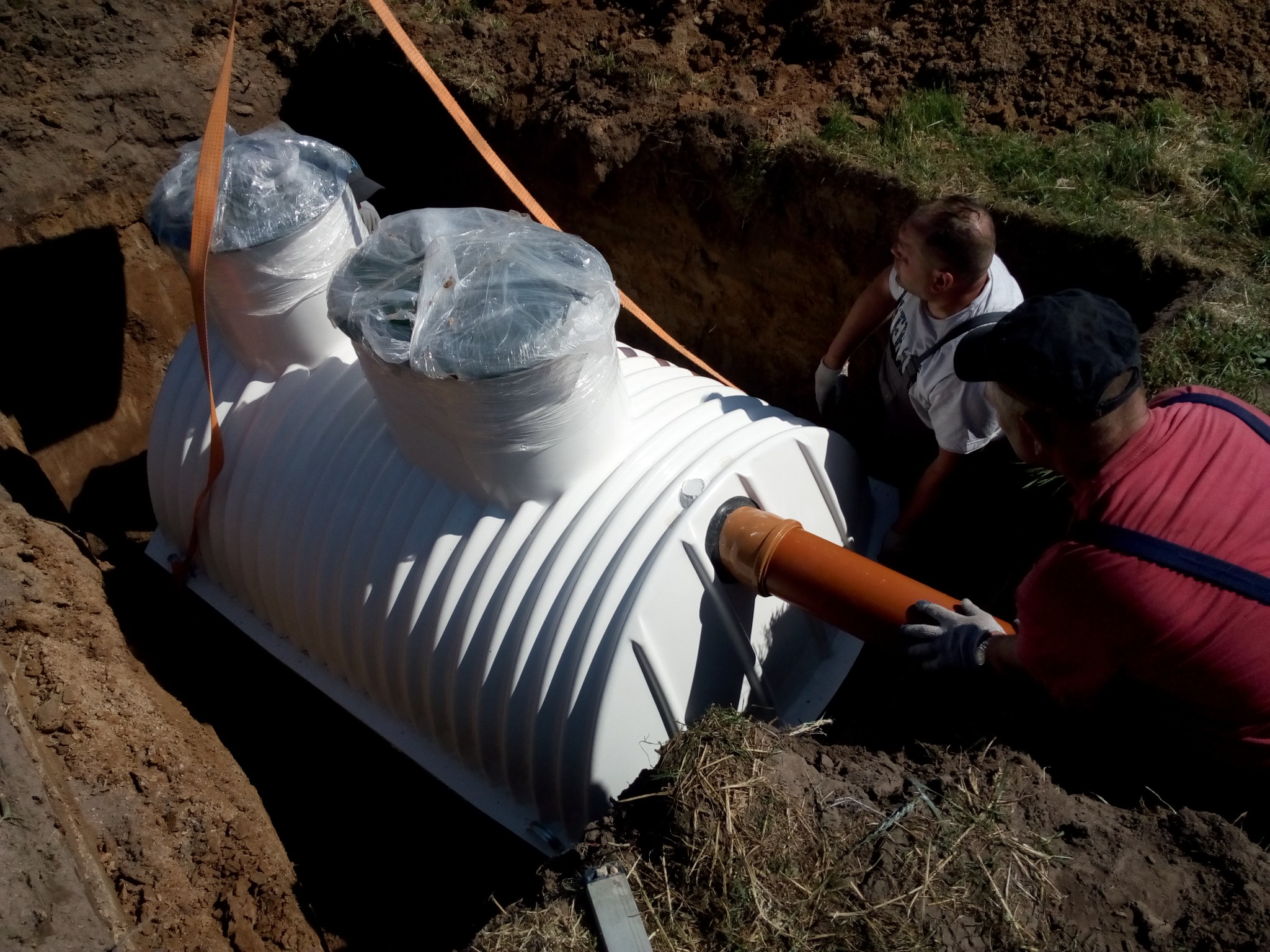 Montaż przydomowej oczyszczalni ścieków: biały zbiornik z tworzywa sztucznego umieszczany w wykopie, pracownicy w roboczych ubraniach podłączają rurę odpływową, zabezpieczone folią wloty.