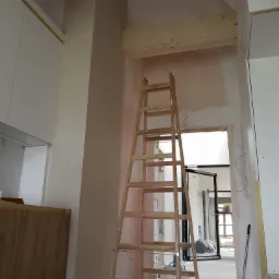 Wnętrze w trakcie prac wykończeniowych: drewniana drabina ustawiona przy ścianie z widocznymi śladami po szpachlowaniu, laserowy niwelator wyznacza poziom. W tle widoczne otwarte drzwi do kolejnego...