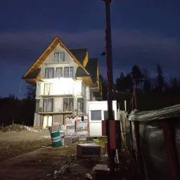 Surowy stan domu w trakcie budowy o zmierzchu, widoczne niezabezpieczone okna, sterty materiałów budowlanych i prowizoryczne oświetlenie placu budowy.