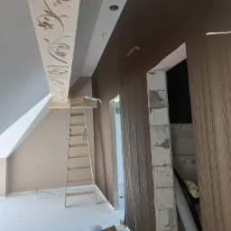 Wnętrze w trakcie prac: drewniana drabina oparta o ścianę, sufit z dekoracyjnym, rzeźbionym elementem, częściowo położona tapeta w pionowe pasy, surowy otwór drzwiowy z widocznym murem z bloczków.