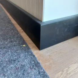 Detale wykończenia wnętrza: czarna listwa przypodłogowa przy białej ścianie i drewnianej podłodze, zabezpieczona szarą włókniną malarską.