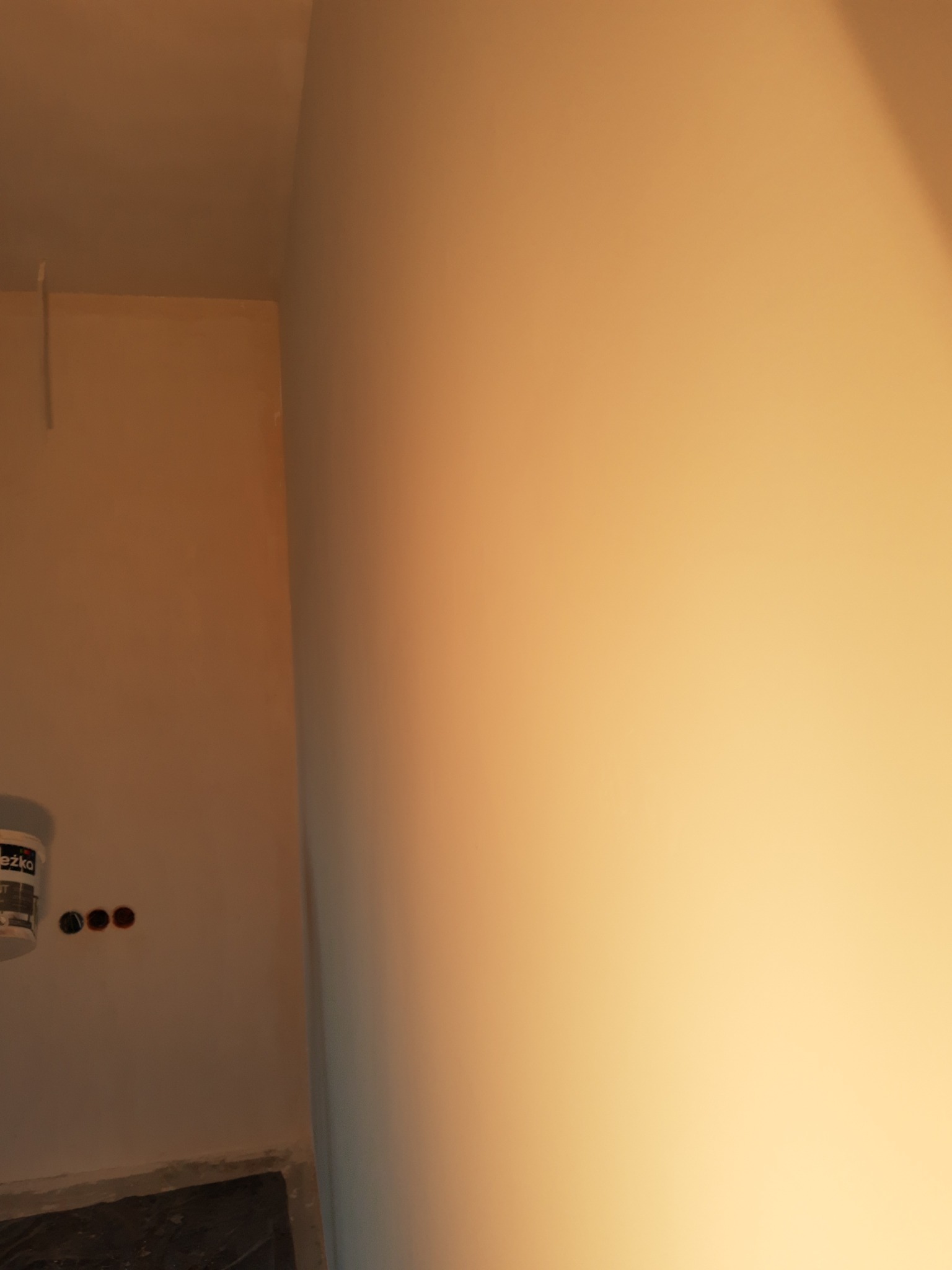 Świeżo pomalowana ściana w rogu pomieszczenia, widoczny fragment puszki z farbą i przygotowane otwory na gniazdka elektryczne.