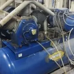 Niebieski, dwucylindrowy kompresor powietrza z widocznym silnikiem elektrycznym i przewodami, ustawiony w warsztacie z zielonymi szafkami w tle.