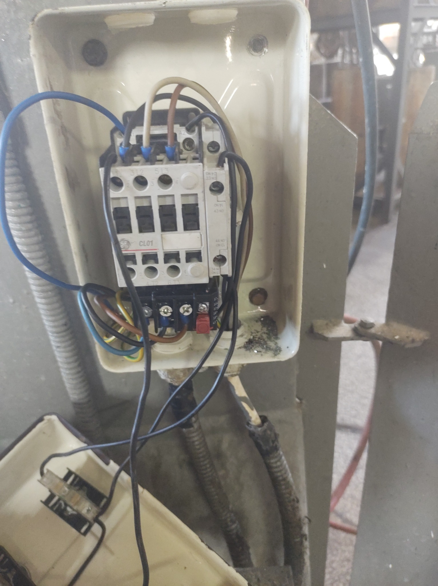 Otwarta skrzynka elektryczna z widocznym stycznikiem CL01 oraz przewodami o różnych kolorach, zamocowana na metalowej konstrukcji. Widoczne ślady zabrudzeń wewnątrz skrzynki.
