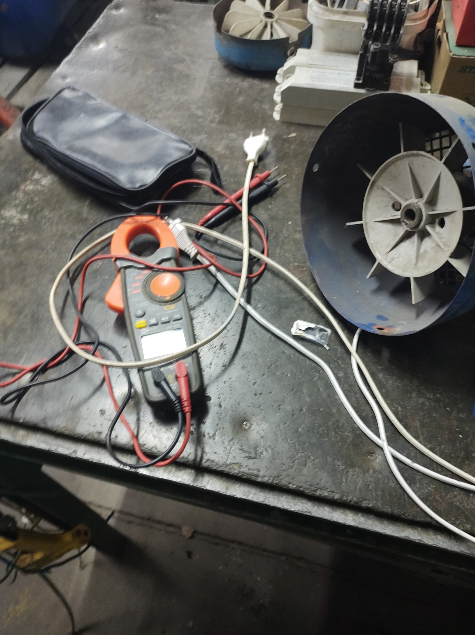 Miernik cęgowy z przewodami pomiarowymi, torba narzędziowa, fragment obudowy wentylatora i inne elementy elektryczne na warsztatowym stole z metalu.