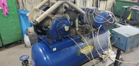 Niebieski, dwucylindrowy kompresor powietrza z widocznym silnikiem elektrycznym i przewodami, ustawiony w warsztacie z zielonymi szafkami w tle.