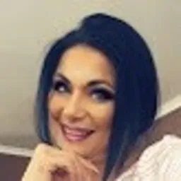 Portret uśmiechniętej kobiety z ciemnymi włosami, opierającej podbródek na dłoni, w jasnym ubraniu, na rozmytym tle.